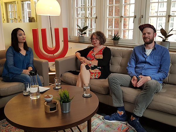 Zwei Frauen und ein Mann sitzen auf Sofas mit dem Logo der Uni Bremen im Hintergrund