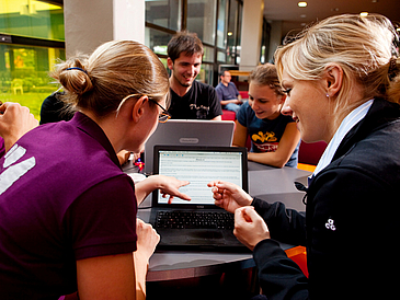 Vordergrund: Zwei Studentinnen diskutieren über einen Text, dargestellt auf einem Laptopmonitor, Hindergrund: Student und Studentin arbeiten an einem Laptop.