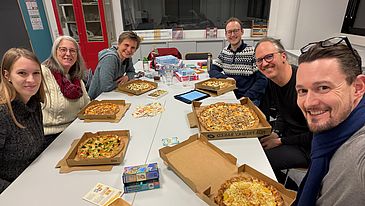 Sechs Personen sitzen um einen Tisch, essen Pizza und spielen gemeinsam Kartenspiele. Auf dem Tisch liegen Pizzakartons und verschiedene Spiele, während alle in die Kamera grinsen. Die Atmosphäre scheint locker und fröhlich.