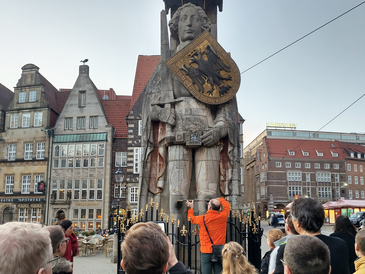 Teilnehmende der mathematischen Stadtführung blicken auf die Statue des Bremer Rolands. Der Stadtführer zeigt dort den Abstand zwischen den beiden Knien.