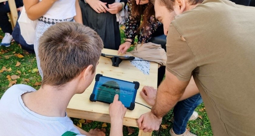Zwei Studenten schauen auf ein Tablet, auf dem eine Spiele-App sichtbar ist. Das Tablet steht auf einem Holztisch, die Studenten sind von hinten zu sehen. Im Hintergrund sieht man mehrere Menschen.