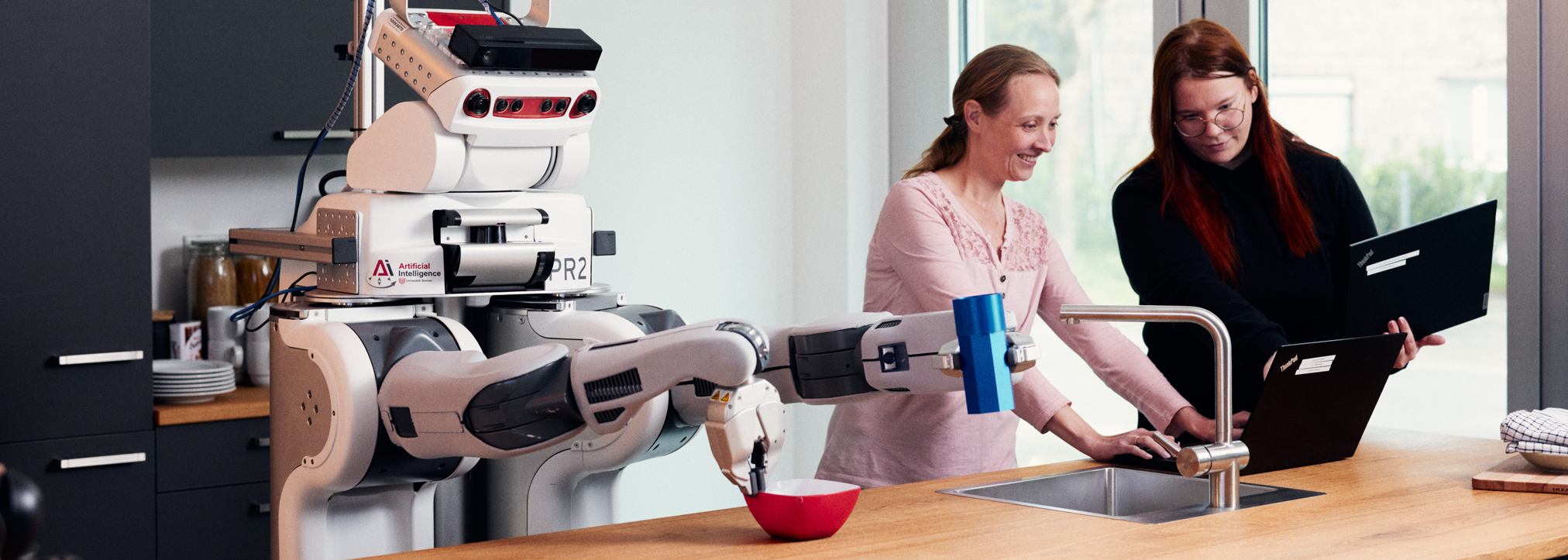 A robot stands behind a kitchen counter together with two women. The women operate a laptop, the robot stirs in a bowl.