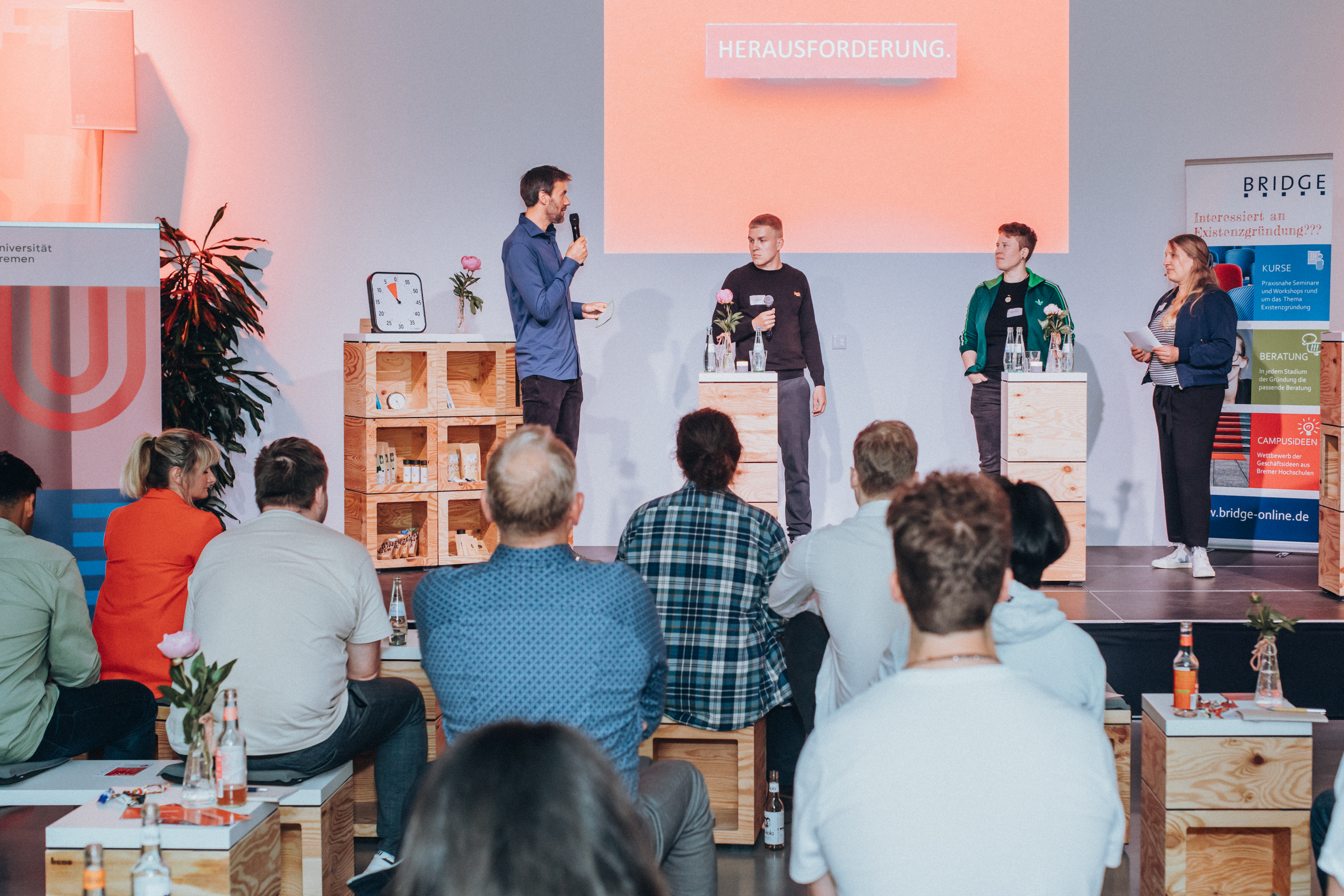 Foto von der StartUp-Lounge: Vier Personen stehen auf einer Bühne, davor sitzt Publikum